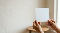 Hands holding blank white card or paper against neutral beige wall background, suitable for mockups, presentations, and advertising campaigns.
