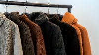 Warm knitted sweaters in earth tones hanging on black rack against white wall. Cozy autumn and winter wardrobe collection featuring brown, orange, and gray colors.