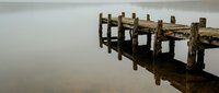 Weathered wooden pier extending into still, misty water with fog obscuring the distant horizon, conveying solitude and tranquility for atmospheric landscape and nature projects.