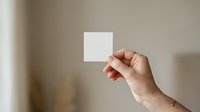 Hand holding blank white square card against neutral beige wall background. Suitable for branding, business card mockups, product packaging design, and marketing presentations.