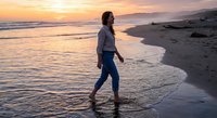 Young woman walking barefoot on sandy beach at golden sunset with ocean waves and peaceful coastal scenery for relaxation and wellness concepts.