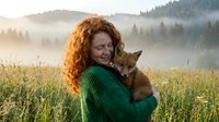 Young woman with curly red hair holding fox cub in green sweater against misty mountain forest landscape at golden hour sunrise creating warm outdoor nature scene.