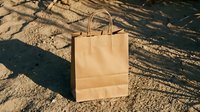 Kraft paper shopping bag with handles sitting on sandy beach with tree shadow overlay, suitable for retail, commerce, and packaging design projects.