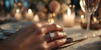 Elegant diamond engagement ring on woman's hand at romantic candlelit dinner table with soft bokeh background.