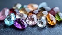 Colorful precious gemstones scattered on dark surface, including blue topaz, amethyst, rose quartz, and citrine crystals in teardrop cut shapes, macro photography.