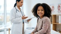 Young African American girl with curly hair smiling during pediatric checkup with female doctor in white coat reviewing medical chart in bright clinic.