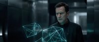Middle-aged man with dark hair and facial stubble, wearing black clothing, interacting with glowing geometric wireframe holographic shapes in a modern tech environment.