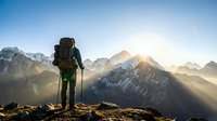Male hiker with backpack standing on mountain peak at sunrise, silhouetted against dramatic alpine landscape with snow-capped peaks and golden sunlight rays.