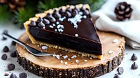 Dark chocolate ganache tart slice on rustic wood serving board garnished with sea salt flakes and chocolate chips, surrounded by pine cones and winter decor.