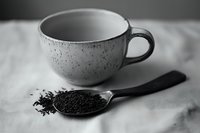 Minimalist black and white composition with ceramic speckled cup and wooden spoon filled with loose tea leaves on light textile background. Perfect for tea packaging.