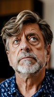 Close-up portrait of mature Caucasian man with gray hair and beard wearing blue patterned shirt, looking up with intense blue eyes and contemplative expression.