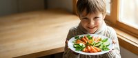 Happy young boy holding colorful healthy meal plate with vegetables and protein in bright kitchen setting for family nutrition and childhood wellness.