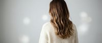 Young woman with long wavy brown hair wearing white knit sweater standing with back turned against wall with glowing light bulbs for creative concepts.