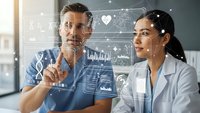 Two diverse medical professionals analyzing futuristic holographic health data interface with charts, DNA strands and vital signs in modern hospital setting.