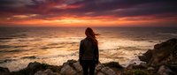 Woman with long brown hair, wearing dark jacket, standing on rocky coastline facing ocean during dramatic sunset with golden and orange sky, contemplative mood, back view, solitude and nature theme.