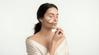 Young woman applying skincare cream with closed eyes and peaceful expression on neutral background for beauty and wellness concepts.