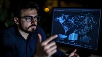 Young Hispanic male data analyst in glasses working late at night, pointing at glowing screen displaying global network visualization and analytics dashboard.