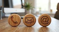 Wooden contact icons on office desk featuring phone, email and at symbol for business communication and customer service concepts.