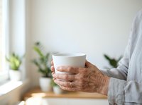 Senior woman with aged hands holding white ceramic mug in bright home interior with green plants, suitable for wellness, lifestyle, and beverage marketing campaigns.