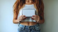 Young woman in crop top and jeans holding blank digital tablet device against light background, showing red manicured nails and casual modern lifestyle.