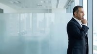 Middle-aged male professional in dark business suit standing by modern office window, contemplating strategy and business decisions in corporate environment.