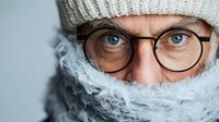 Close up portrait of mature Caucasian man wearing glasses and winter white knit hat, face partially covered with warm scarf, looking directly at camera with blue eyes.