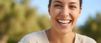 Happy young Hispanic woman with bright smile laughing outdoors on sunny day with natural green background for wellness and lifestyle content.