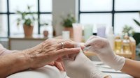 Healthcare worker in white gloves gently holding elderly patient hands during medical examination in bright clinical room with plants and natural light.