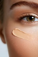 Close-up of flawless skin with foundation cream streak on cheek, brown eye with long lashes and defined eyebrow showing professional makeup application.