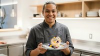 Young male chef of mixed ethnicity wearing dark chef's coat, smiling while holding plate with gourmet plated dish in modern kitchen with open shelving, suitable for culinary, restaurant, and food service marketing.