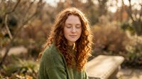 Young woman with curly red hair meditating peacefully in golden sunlight outdoors, eyes closed in serene contemplation for wellness and mindfulness concepts.