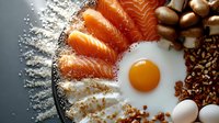 Overhead view of breakfast ingredients arranged in sections: smoked salmon slices, fried egg yolk, coffee beans, nuts, flour powder, and eggs on gray surface for culinary and food styling projects.