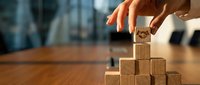 Person with medium skin tone placing wooden block on pyramid stack, representing business growth, strategy and success planning on wooden desk.