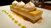 Layered citrus dessert with meringue topping on white plate, garnished with lime sauce dots and dusted cinnamon, served at upscale restaurant setting.