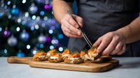 Chef preparing gourmet appetizers on wooden board with festive Christmas lights creating elegant holiday dining atmosphere for celebration.