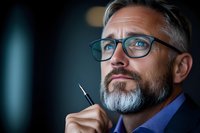 Thoughtful mature Caucasian businessman with gray beard and glasses holding pen while contemplating important decision against dark background. Professional portrait.