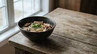 Steaming bowl of Asian noodle soup with vegetables and herbs served in black ceramic bowl on rustic wooden table near window for cozy dining atmosphere.