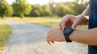 Male runner checking smartwatch during outdoor workout on sunny day in park setting with blurred green trees and path background.