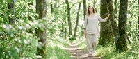 Middle-aged Caucasian woman in casual beige clothing walking on forest trail surrounded by green trees and natural woodland scenery for lifestyle and wellness content.