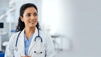 Professional Hispanic female doctor in white coat with stethoscope smiling confidently in modern medical clinic setting for healthcare marketing.