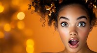Surprised young African American woman with curly hair and wide eyes against warm golden bokeh lights background for holiday celebrations.