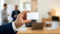 Male professional in business attire holding blank white card in modern office environment with colleagues in background. Suitable for corporate branding and networking concepts.