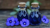 Two small glass bottles with cork stoppers filled with deep blue liquid surrounded by purple pansy flowers on rustic wooden surface for aromatherapy and wellness.