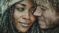 Intimate winter couple portrait with snowflakes, young mixed-race woman and bearded Caucasian man sharing tender moment in cold weather outdoors.