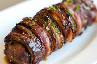 Succulent bacon-wrapped pork tenderloin sliced and garnished with fresh herbs, glazed with sweet and savory sauce, served on white plate for restaurant menu.