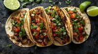 Mexican street tacos with shredded meat topped with fresh cilantro, diced tomatoes, and onions on corn tortillas, served with lime wedges on dark rustic background.