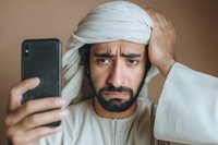 Middle Eastern man in traditional white headwear looking confused at smartphone screen with hand on forehead expressing frustration and technology stress.
