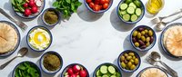 Fresh Mediterranean ingredients arranged in colorful bowls on white marble surface with olive oil, herbs, and vegetables for healthy cooking preparation.