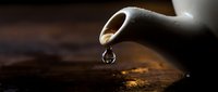 Ceramic teapot spout with a single water drop falling onto a dark wooden surface, captured in dramatic close-up macro photography.