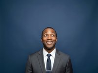 Smiling male professional with dark skin tone wearing gray suit and navy tie against solid blue background, suitable for corporate portraits and business headshots.